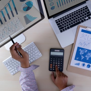 sage accounting software price cost man on pocket calculator with pen in left hand laptop and monitor on desk white wireless keyboard